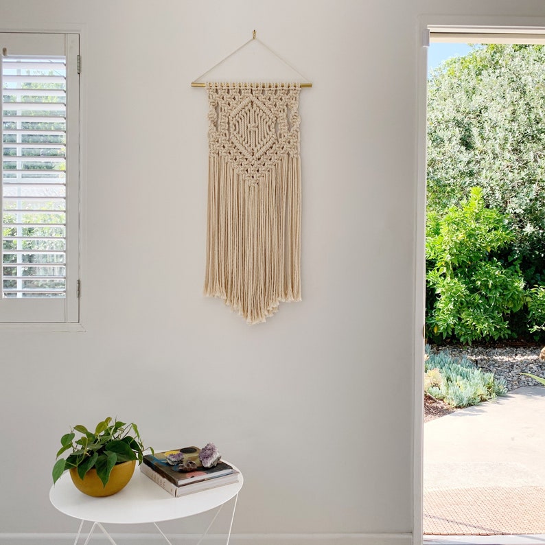 May include: A white macrame wall hanging with a geometric pattern, hanging from a gold rod on a white wall. The wall hanging is in a room with a white window and a door leading to a patio.