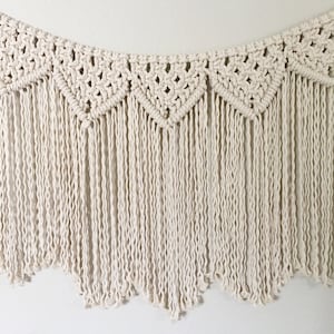 May include: A white macrame wall hanging with a scalloped edge and fringe. The hanging is made of cotton cord and has a geometric pattern.