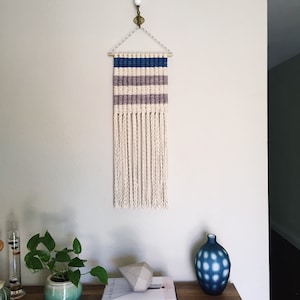 Może przedstawiać: Wiszący gobelin tkany z białym tłem i niebieskimi i fioletowymi paskami. Gobelin ma frędzle z białej przędzy.