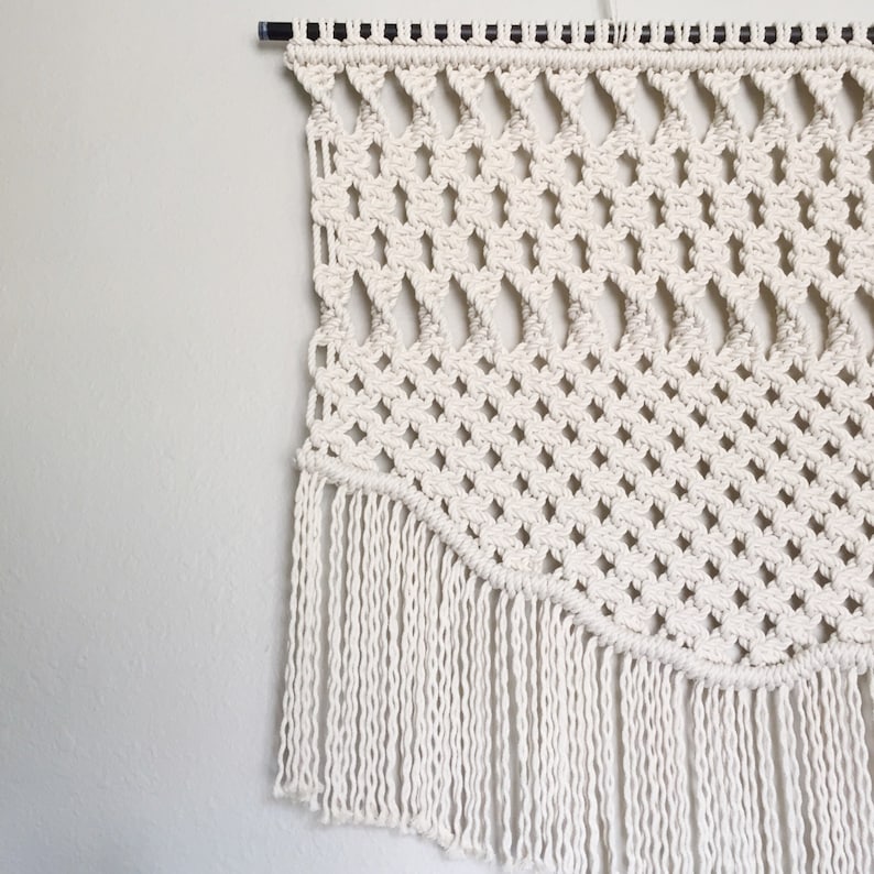May include: Cream-colored macrame wall hanging with a geometric diamond pattern and fringe. The wall decor is suspended from a black rod and is displayed against a white wall. The macrame design adds a bohemian touch to any space.