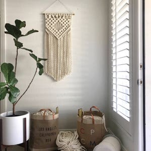 May include: A white macrame wall hanging with a geometric pattern, two brown canvas tote bags with the text "Los Angeles California USA" and "CP CM" printed on them, and white yarn balls.