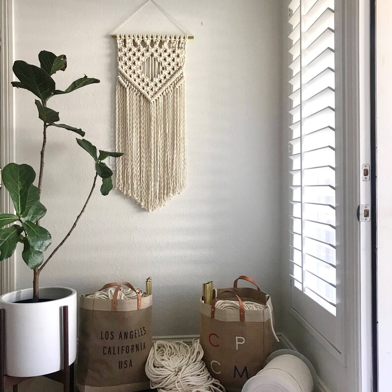 May include: A white macrame wall hanging with a geometric pattern, two brown canvas tote bags with the text "Los Angeles California USA" and "CP CM" printed on them, and white yarn balls.