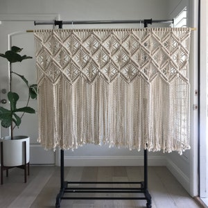 May include: A white macrame wall hanging with a geometric pattern and fringe. The hanging is displayed on a black metal stand.