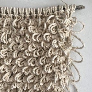 May include: A large, cream-colored macrame wall hanging with a natural wood branch at the top. The macrame is made with thick, knotted rope and has a textured, organic look.
