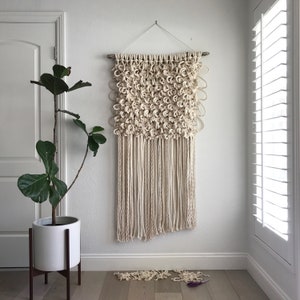 May include: A white macrame wall hanging with a textured, knotted design. The hanging is made of natural fibers and has a fringe at the bottom.