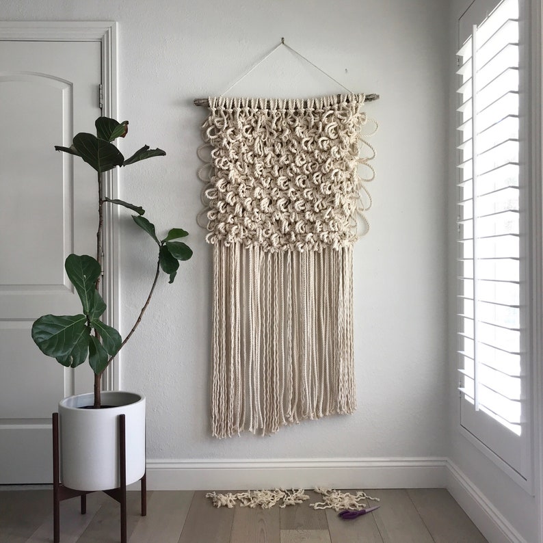 May include: A white macrame wall hanging with a textured, knotted design. The hanging is made of natural fibers and has a fringe at the bottom.