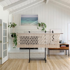 May include: A white macrame wall hanging with a diamond pattern and fringe, hanging on a black metal stand in front of a white brick fireplace.