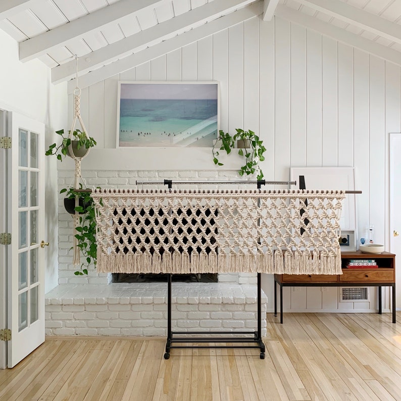 May include: A white macrame wall hanging with a diamond pattern and fringe, hanging on a black metal stand in front of a white brick fireplace.