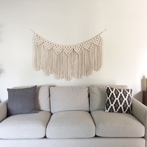May include: A white macrame wall hanging with a scalloped edge hangs above a light grey sofa with two grey throw cushions and one black and white patterned throw cushion.