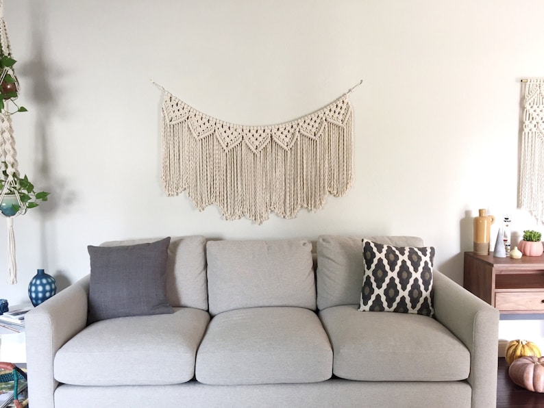 May include: A white macrame wall hanging with a scalloped edge hangs above a light grey sofa with two grey throw cushions and one black and white patterned throw cushion.