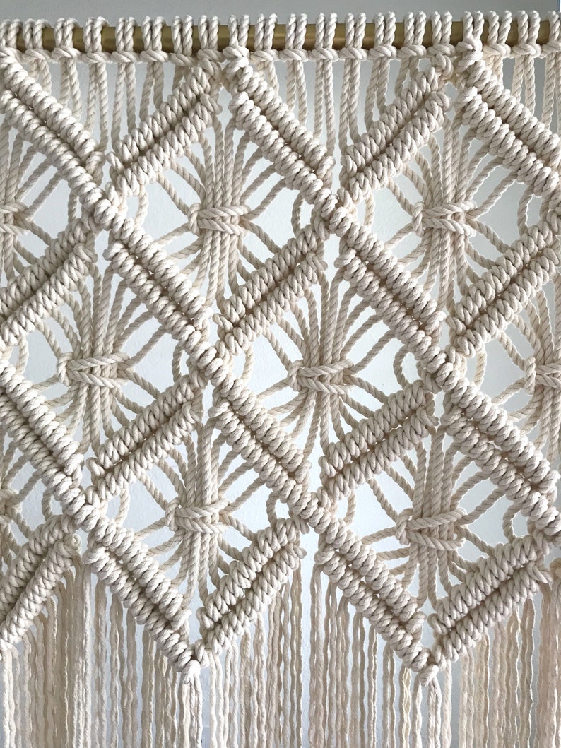 Puede incluir: Un tapiz de macram&eacute; blanco con un patr&oacute;n geom&eacute;trico de diamantes y flecos. El tapiz est&aacute; hecho con cord&oacute;n de algod&oacute;n natural y est&aacute; colgado de una varilla dorada.