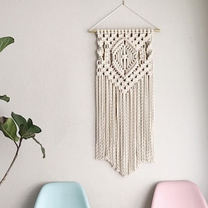 May include: A white macrame wall hanging with a geometric design and fringe. The hanging is attached to a gold rod.