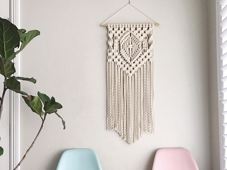 May include: A white macrame wall hanging with a geometric design and fringe. The hanging is attached to a gold rod.