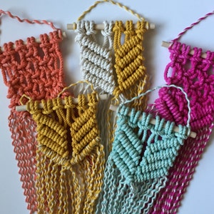 May include: Five macrame wall hangings with wooden dowels. The hangings are in various colors, including coral, yellow, white, teal, and pink.