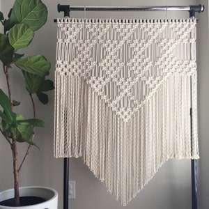May include: A large, white macrame wall hanging with a geometric pattern and fringe. The hanging is displayed on a black stand against a white wall.