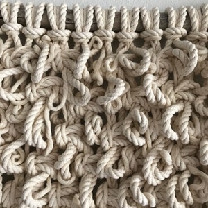 May include: A close-up of a macrame wall hanging made with natural colored rope. The rope is knotted in a complex pattern, creating a textured and organic look.
