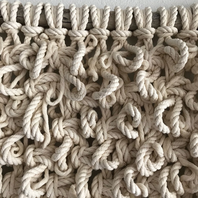 May include: A close-up of a macrame wall hanging made with natural colored rope. The rope is knotted in a complex pattern, creating a textured and organic look.