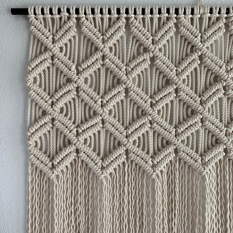 May include: A macrame wall hanging with a geometric diamond pattern in a cream color. The hanging is made with thick cotton cord and has a fringe at the bottom.