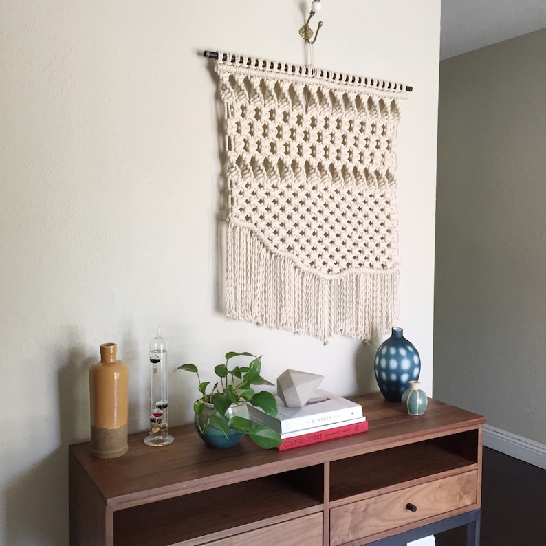 May include: A cream-colored macrame wall hanging with a geometric design and fringe detail. The wall decor is suspended from a black rod and a decorative hook. A wooden console table displays decorative vases, books, and a small potted plant.