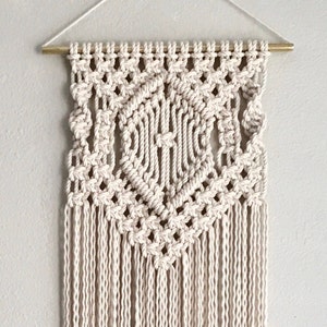 May include: A white macrame wall hanging with a geometric diamond pattern and long fringe. The hanging is made with natural cotton cord and is attached to a wooden dowel.
