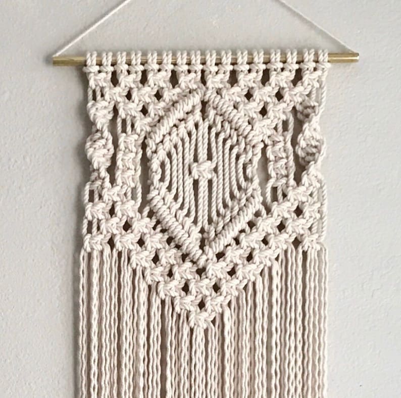May include: A white macrame wall hanging with a geometric diamond pattern and long fringe. The hanging is made with natural cotton cord and is attached to a wooden dowel.