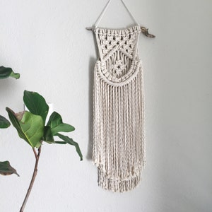 May include: A white macrame wall hanging with a geometric design and fringe. The hanging is attached to a natural branch.