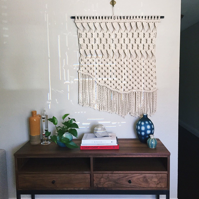 May include: A cream-colored macrame wall hanging with a geometric pattern and fringe detail. Below, a dark wood console table displays decorative vases, books, and a potted plant. The wall is painted a light gray.