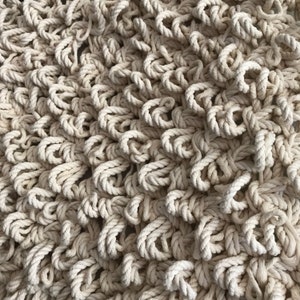 May include: A close-up of a cream-colored macrame wall hanging made from thick rope. The rope is woven into a pattern of loops and knots, creating a textured and organic look.
