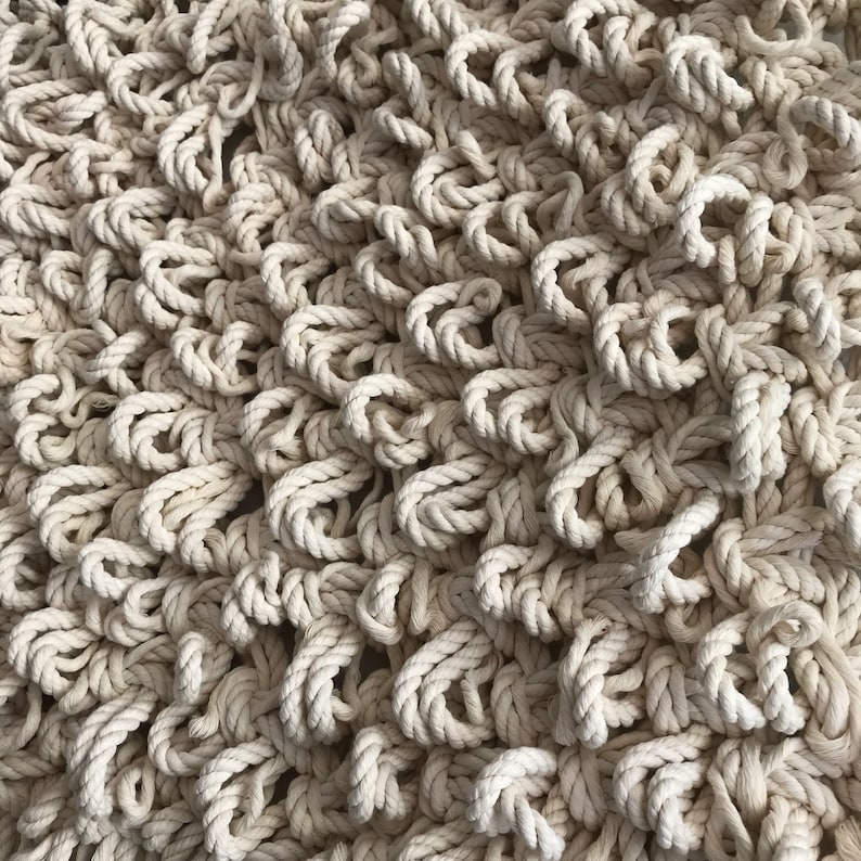 May include: A close-up of a cream-colored macrame wall hanging made from thick rope. The rope is woven into a pattern of loops and knots, creating a textured and organic look.