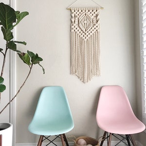 May include: Two chairs, one light blue and one pink, with a macrame wall hanging in the background. The chairs are wooden with a woven basket in front of them.