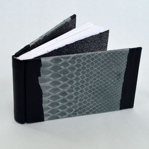 May include: A small, black, leather-bound book with a gray snakeskin pattern on the cover. The book is open to reveal white pages.