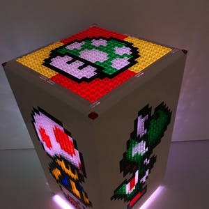 May include: A white cube-shaped lamp with a light-up base. The sides of the cube feature pixelated images of characters from the Super Mario Bros. video game series, including a green Yoshi, a red and white Toad, and a green mushroom.