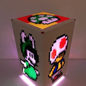 May include: A white cube-shaped lamp with a light pink glow. The sides of the cube feature pixelated images of a green mushroom, a green character with a white hat, and a red and white mushroom character.