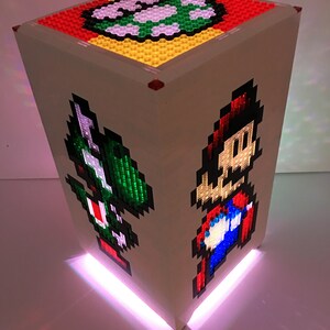 May include: A white cube-shaped lamp with a pixelated image of Mario and Luigi on the sides. The top of the cube has a pixelated image of a Super Mario Bros. power-up. The lamp is lit with a pink light.