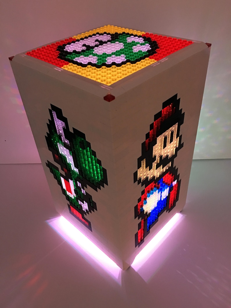 May include: A white cube-shaped lamp with a pixelated image of Mario and Luigi on the sides. The top of the cube has a pixelated image of a Super Mario Bros. power-up. The lamp is lit with a pink light.