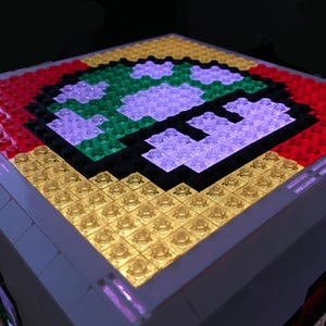 May include: A close-up of a light-up Lego model of a Super Mario mushroom. The mushroom is made of red, green, purple, and yellow Lego bricks. The model is lit from the inside, creating a glowing effect.