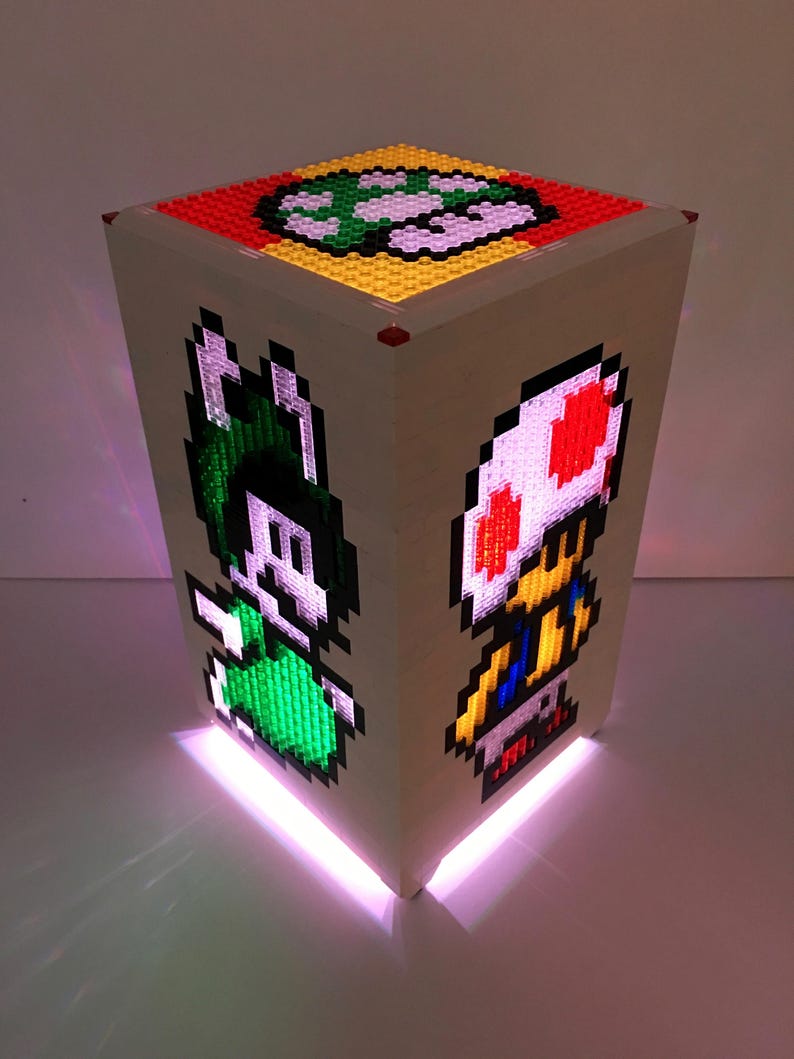 May include: A white cube-shaped lamp with a light pink glow. The sides of the cube feature pixelated images of a green mushroom, a green character with a white hat, and a red and white mushroom character.