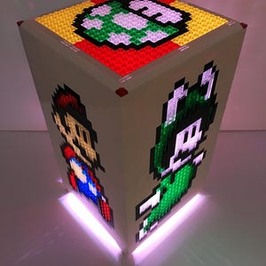 May include: A white Lego cube with a light inside. The cube has a green, yellow, and red design on the top, and images of Mario and Luigi on the sides. The light shines through the Lego bricks, creating a colorful glow.