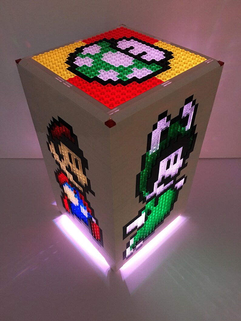 May include: A white Lego cube with a light inside. The cube has a green, yellow, and red design on the top, and images of Mario and Luigi on the sides. The light shines through the Lego bricks, creating a colorful glow.