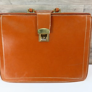 May include: A vintage brown leather briefcase with a rectangular shape. The briefcase features a gold-colored clasp and handle hardware. White stitching outlines the edges of the briefcase. The briefcase is closed and sits on a white surface.