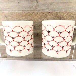 May include: Two cream-colored ceramic mugs with a repeating pattern of red semi-circles and black triangles. Each mug has a white handle. The mugs are displayed on a clear shelf against a wooden background.
