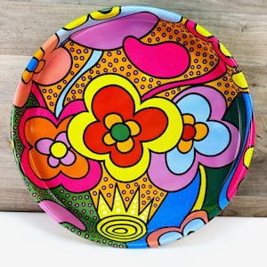 May include: A round, colorful tray with a vibrant floral design. The tray features large, stylized flowers in shades of red, yellow, pink, and blue, set against a background of orange and green. The tray's rim is a deep green.