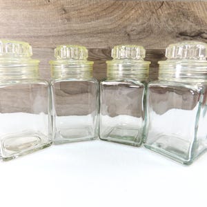 May include: Four clear glass jars with square bodies and yellow-tinted lids. The jars are empty and sit on a white surface against a wooden background. The lids have a decorative, ridged design.