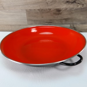 May include: A round, red enamel pan with a silver rim and a black handle. The pan is empty and sits on a white surface, with a wooden background.