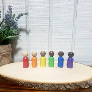 May include: Seven wooden peg dolls in a rainbow color pattern. The dolls are arranged in a line on a wooden slice. The dolls are painted red, orange, yellow, green, blue, purple, and brown.