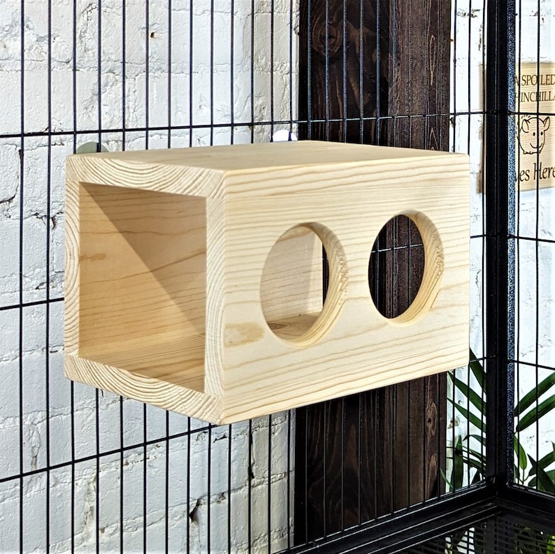 May include: Wooden cage accessory with two round holes and a square opening. The accessory is made of natural wood and is attached to a black wire cage.