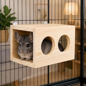May include: A light-colored wooden box hideout for small animals, featuring a large open front and two circular side cutouts. A grey chinchilla is positioned inside the wooden structure, which is mounted to the black wire bars of a cage.
