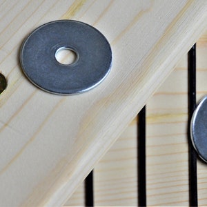 May include: Close-up of hardware components, including a silver screw, a nut, and a washer, arranged on a light-coloured wooden surface. The components are for assembly or repair, likely for furniture or a similar project.