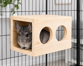 May include: A light-coloured wooden hideout box for small animals, featuring a large rectangular opening on one side and two circular cutouts on the front panel. A grey chinchilla is positioned inside the wooden structure, which is mounted to a black wire cage.