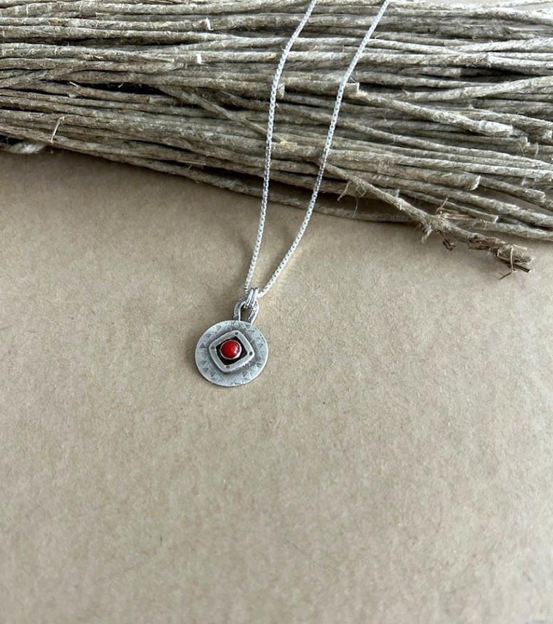 May include: A silver necklace featuring a circular pendant with a hammered texture and a central square frame containing a small, round, vibrant red stone. The pendant hangs from a delicate silver box chain, showcasing a polished metallic finish.